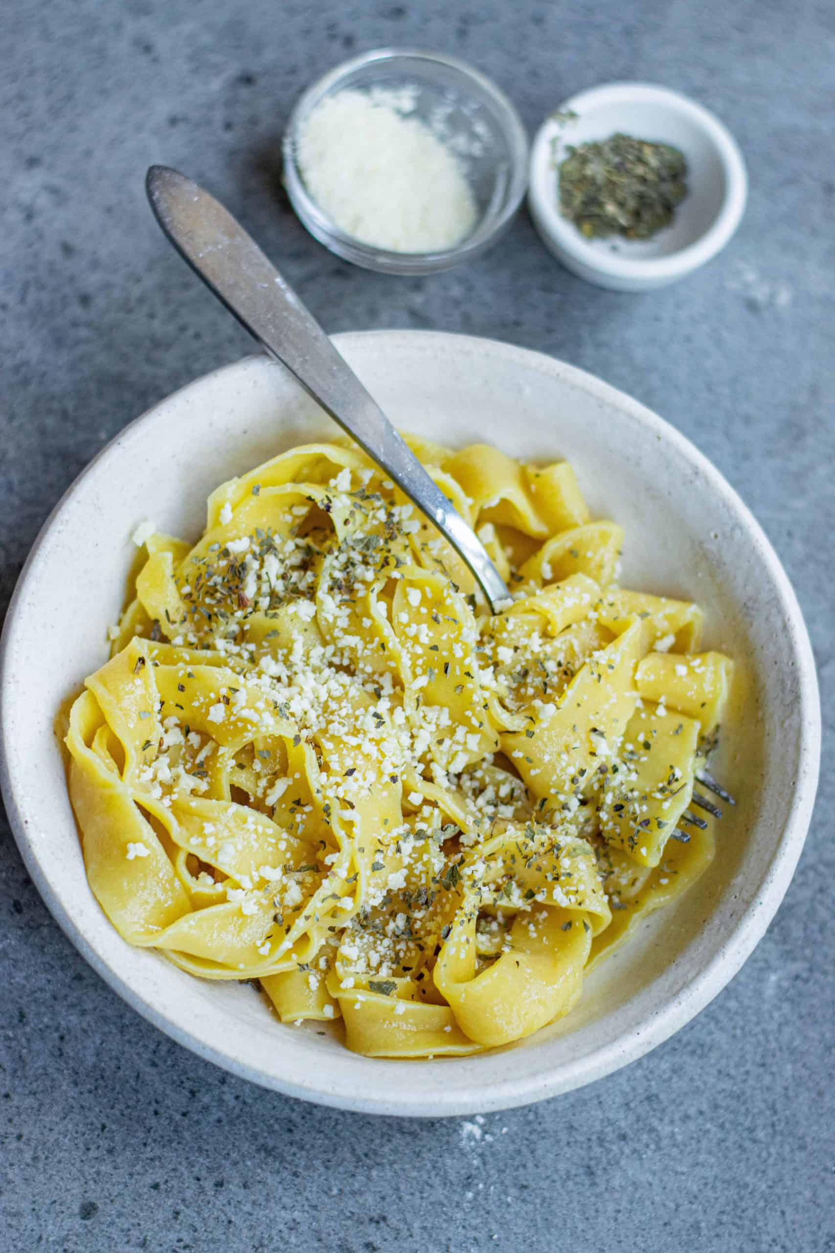 Homemade pumpkin pasta in a white bowl with a fork in it with cheese sprinkled on top. Salt and pepper in small bowls above the pasta.