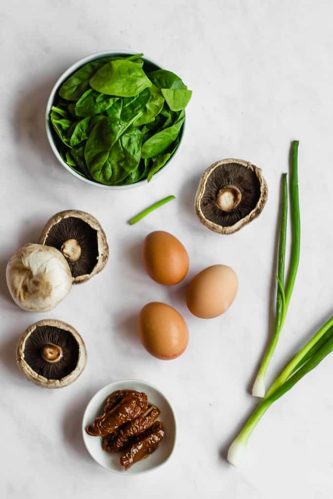 Spinach in a bowl, sun dried tomatoes in a bowl, 4 whole mushrooms, 3 eggs and 2 green onions on a white marble background.