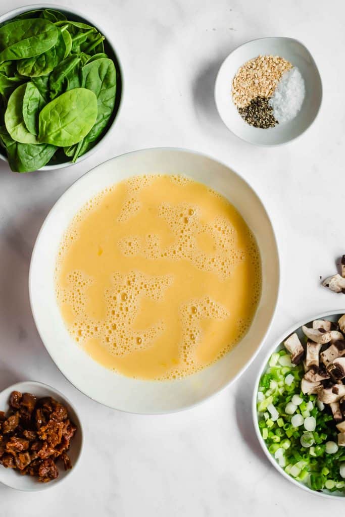 Spinach in a bowl, eggs whipped in a white bowl, bowl of spices, bowl of chopped sound dried tomatoes and a bowl of diced green onions and mushrooms on a white marble background.