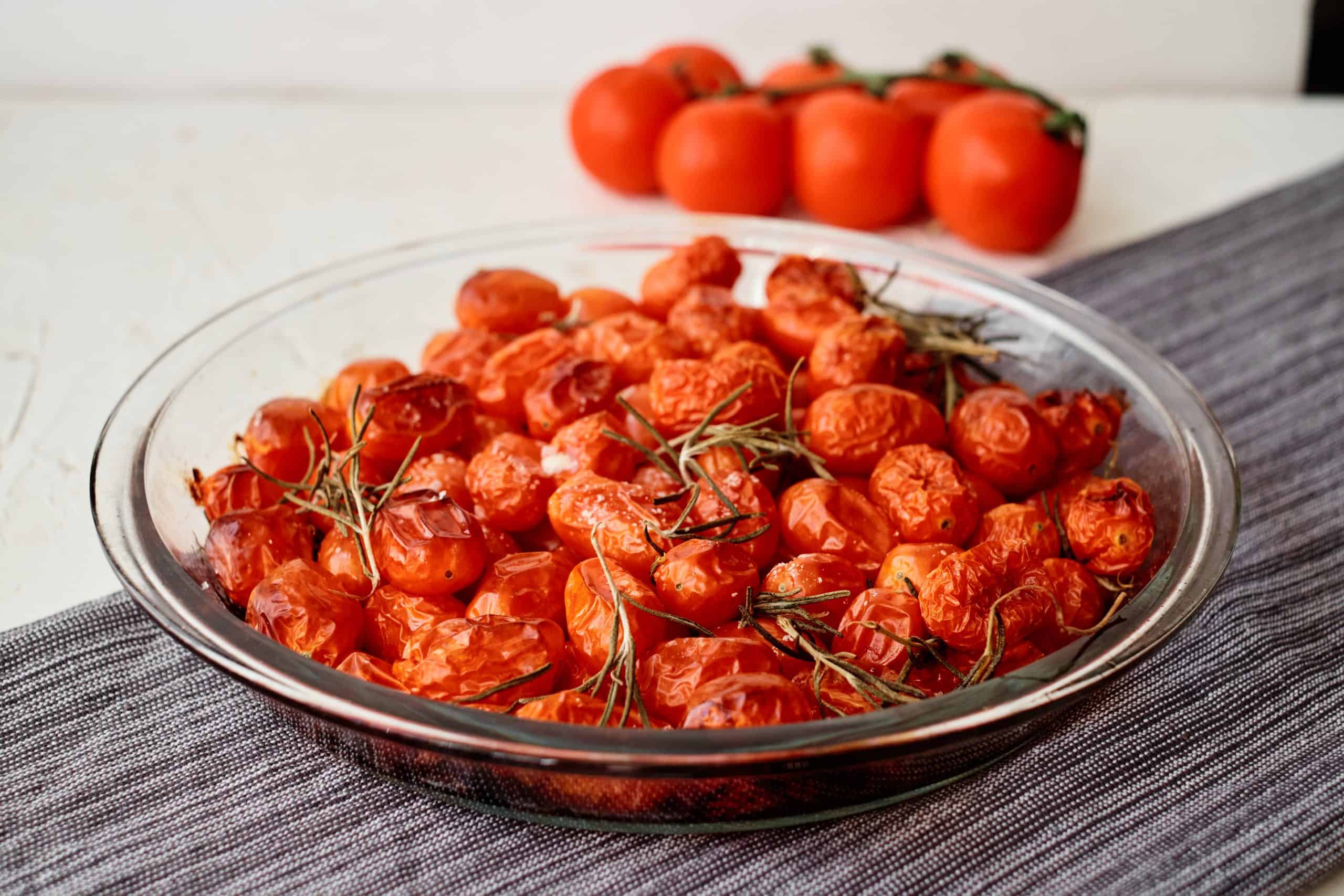 Cherry tomatoes with sprigs of fresh rosemary in a glass pie dish with additional tomatoes behind it.
