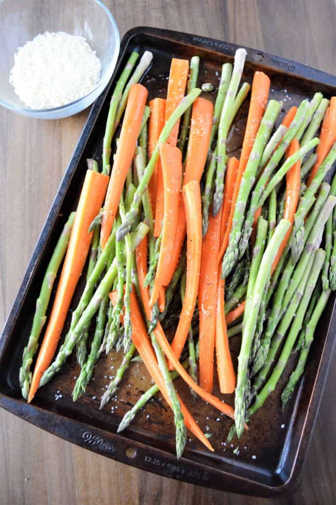 Carrots and asparagus on a baking sheet with Parmesan cheese in a glass bowl in the upper corner.