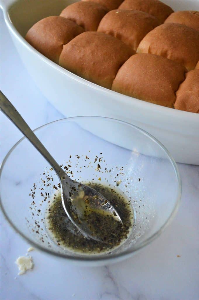 Butter and herb sauce in a glass bowl beside rolls in a white dish.