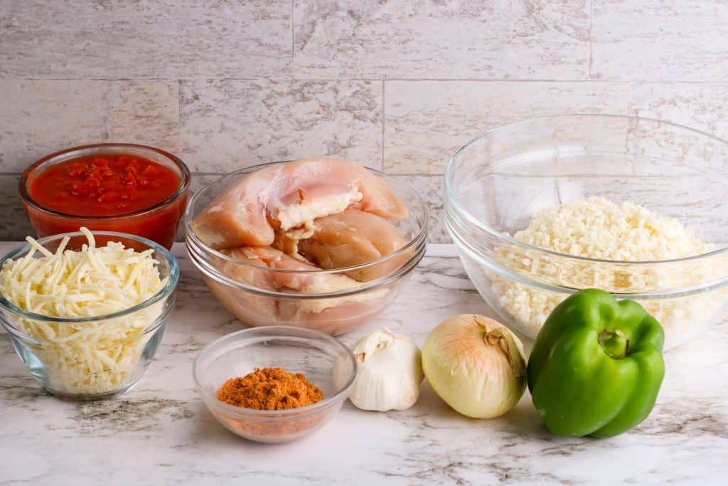Chicken, diced tomatoes, cheese, cauliflower rice, taco seasoning, bell pepper, onion and garlic on a marble counter top.