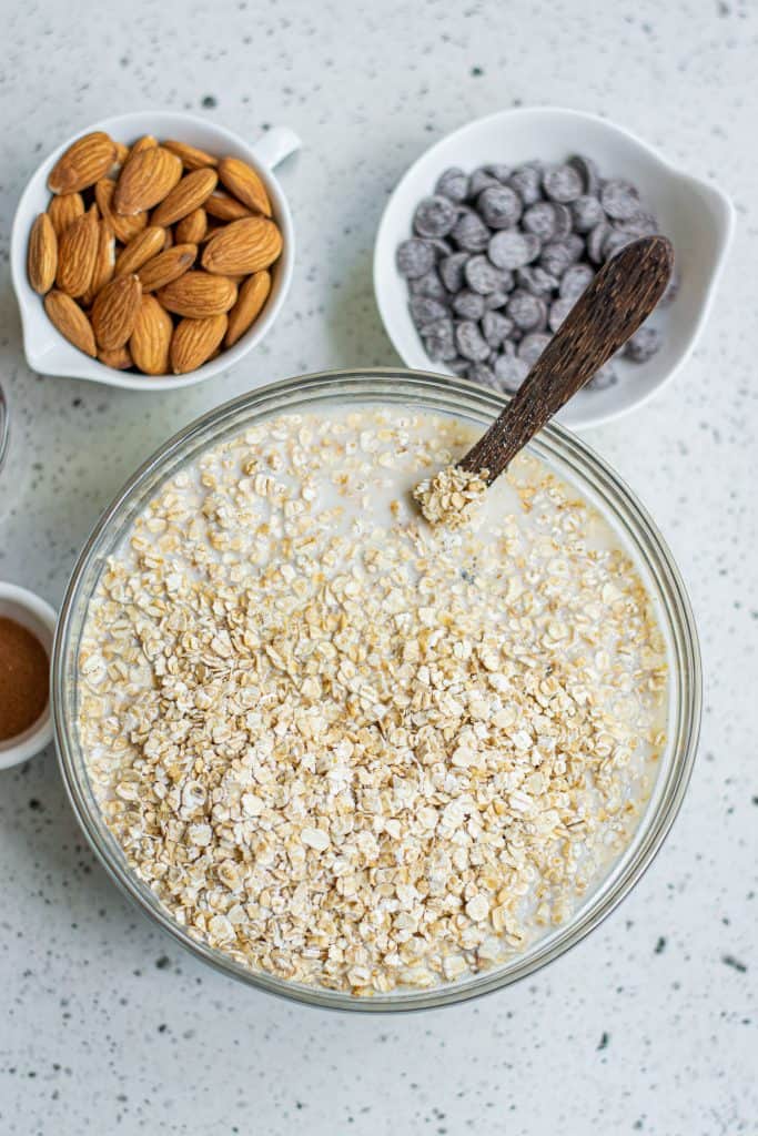 Oats in a glass mixing bowl with a wooden spoon with chocolate chips and almonds in small bowls above it and dried cranberries to left of the bowl.