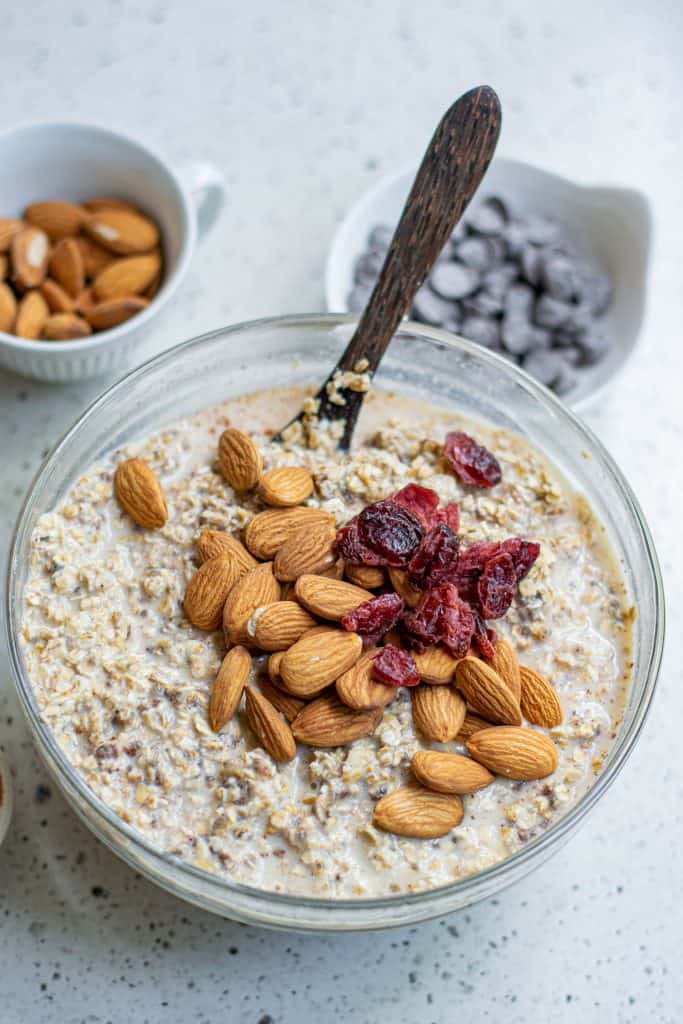 Almonds and dried cranberries on top of oats in a large glass mixing bowl.
