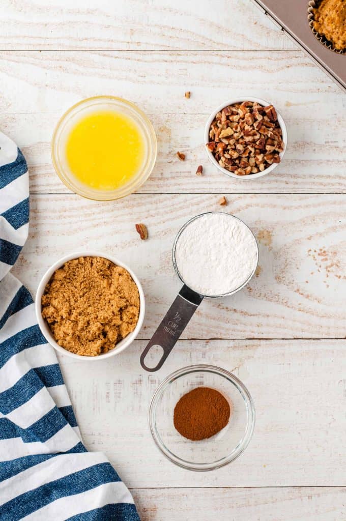 Melted butter in a glass bowl, brown sugar in a white bowl, flour in a measuring cup, cinnamon in a glass bowl and pecans in a white bowl on a white wood background.