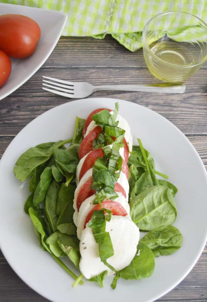 Caprese Salad with Spinach on a white plate with a fork above the plate and a glass of white wine on one side and Roma tomatoes on the other side on a wood background.