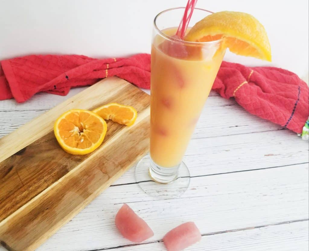 Tall glass with orange juice and orange wedge in it. Cranberry ice cubes in front of it and orange slices on a cutting board behind it.