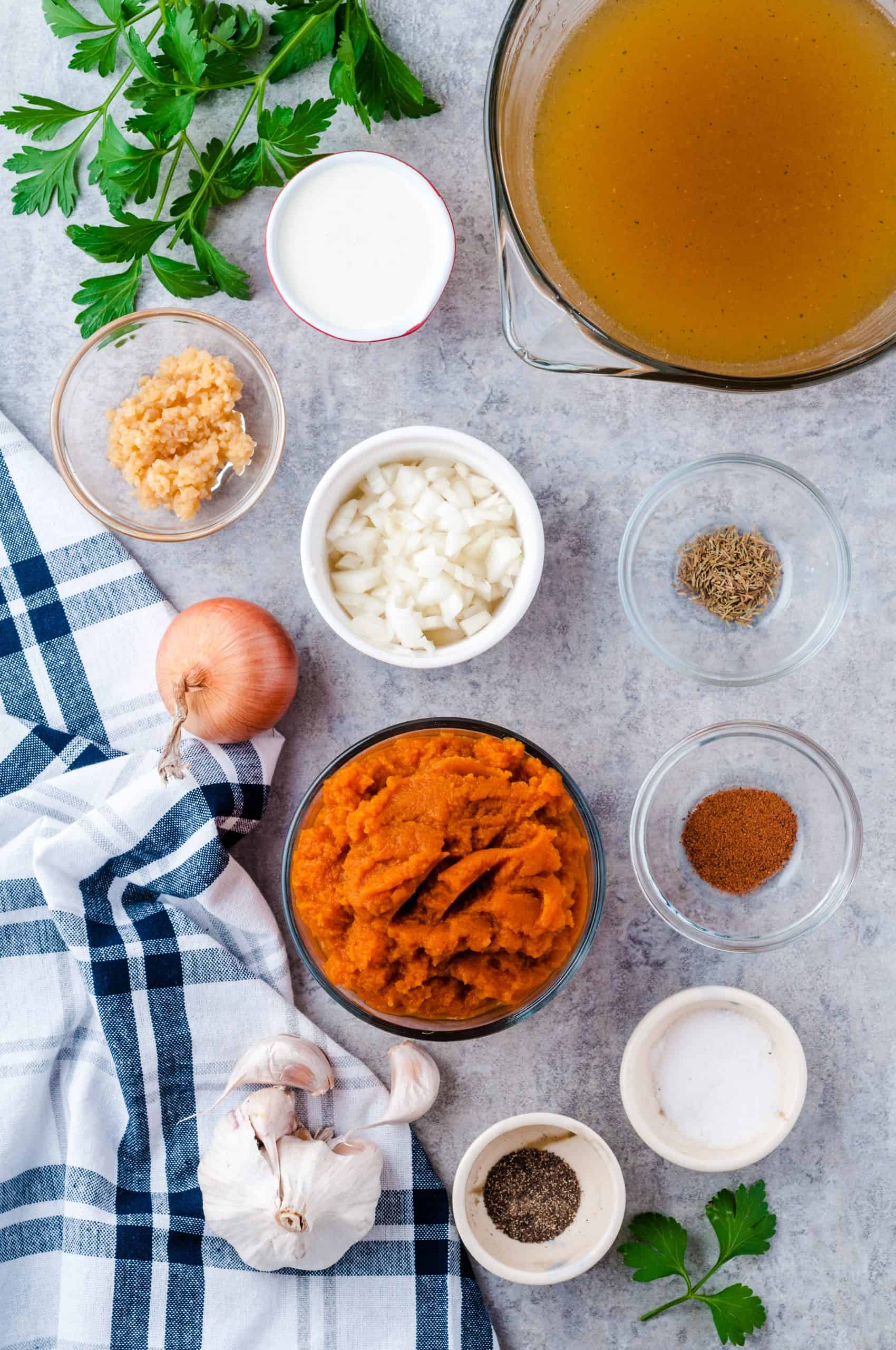 Ingredients for vegetarian pumpkin soup on a gray countertop with a white and blue checked towel on the left side.