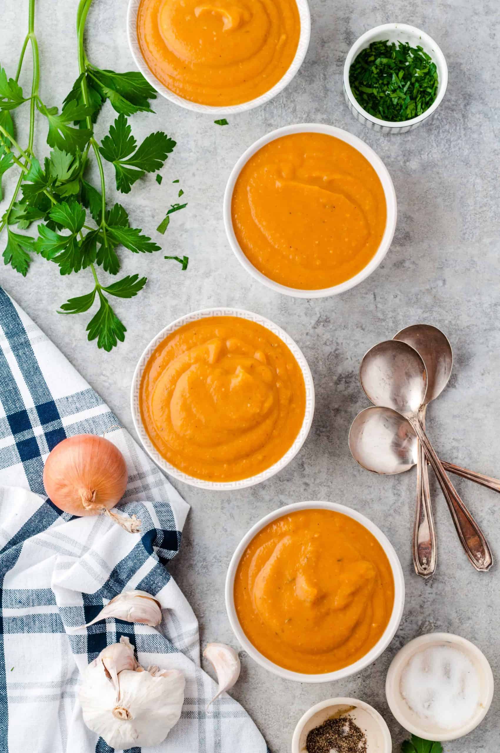 4 servings of pumpkin soup in white bowls with toppings on the side.