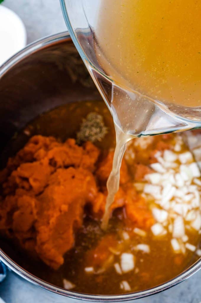 Vegetable broth being poured into pot with pumpkin and onions.