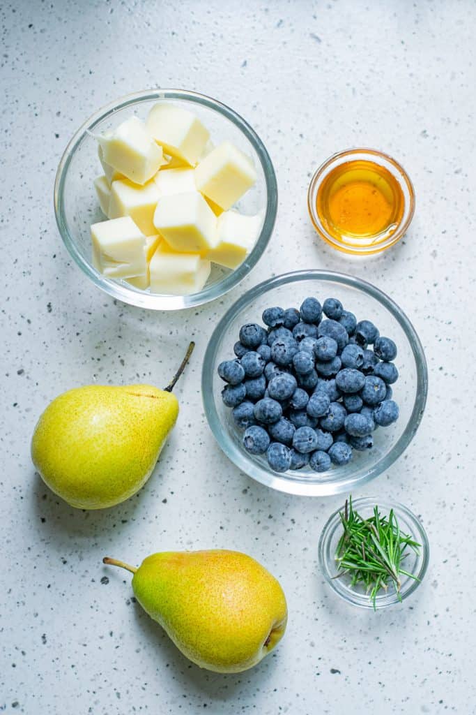 Pears, blueberries, honey, mozzarella cheese and fresh rosemary on a white countertop.