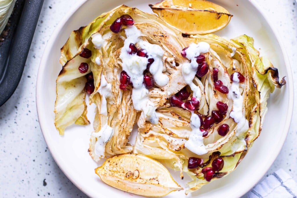 Overhead shot of roasted cabbage with lemon wedges, tartar sauce and pomegranate seeds in a white bowl.