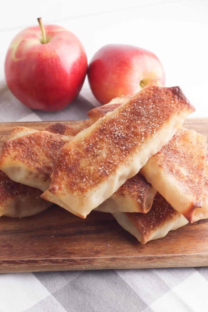 Apple pie egg rolls on a wood cutting board on top of a grey checkered table cloth with whole apples behind it.