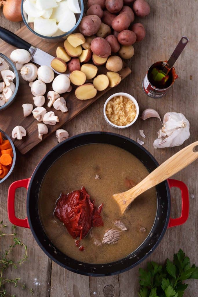 tomato paste on top of beef brith with onions mushrooms, carrots and potatoes on a cutting board above it.
