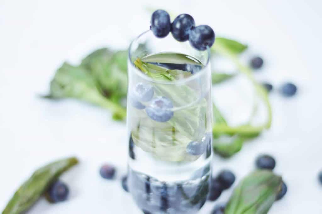 Water glass with blueberries on the rim and blueberries floating in the glass with fresh leaves of basil sprinkle around the glass on a white background.
