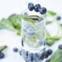 Water glass with blueberries on the rim and blueberries floating in the glass with fresh leaves of basil sprinkle around the glass on a white background.