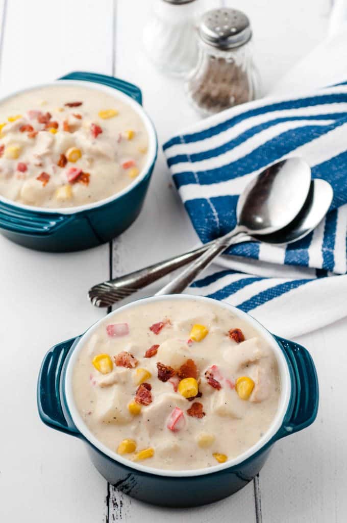 Angled view of corn chowder in blue enamel bowls with 2 spoons on a blue and white striped dish towel on a white wood bacground.
