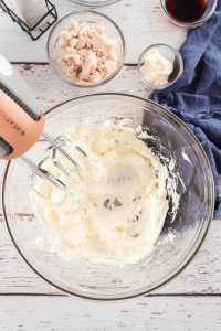A glass mixing bowl with cream cheese blended with a hand mixer.