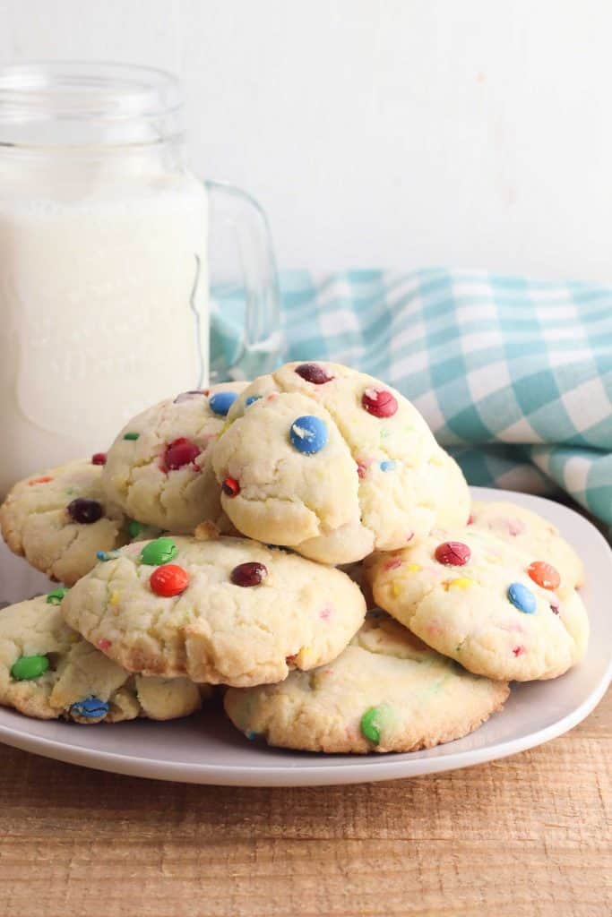 Angled shot of gluten free Funfetti cookies on a wooden table with mason jar all of milk behind it and a light blue and white checked napkin behind the milk and cookies.