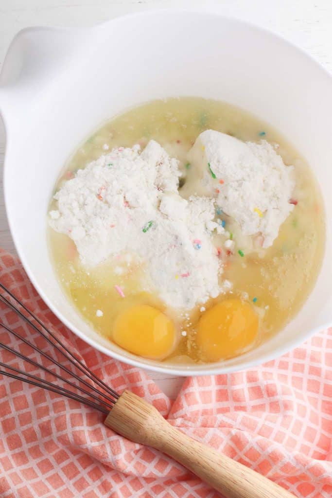 Overhead view of cake mix, eggs and oil in a white bowl with a whisk on the side.