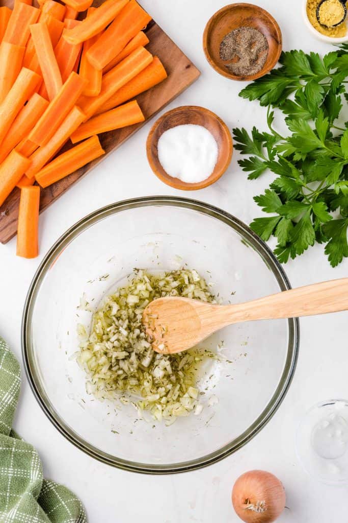 Clear glass bowl with onions, olive oil and herbs in it with a wooden spoon in the bowl and carrots, parsley and salt on the side.