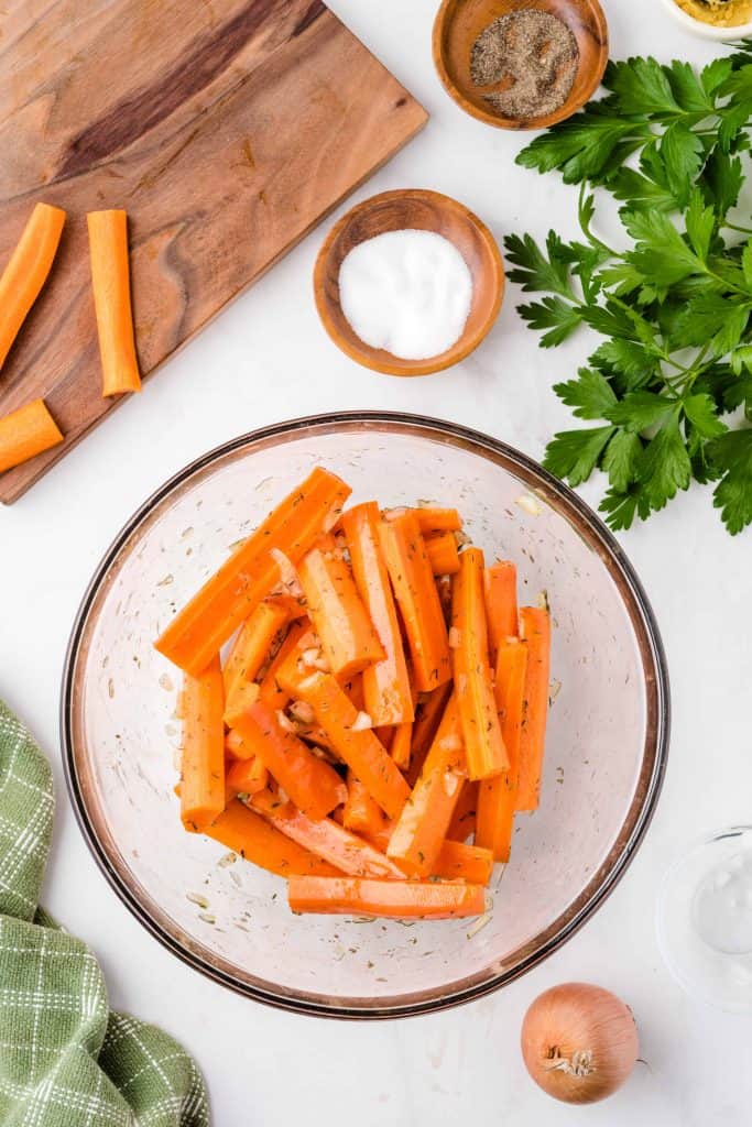 Carrots in a large glass bowl dressed with olive oil and seasoning on a white background.
