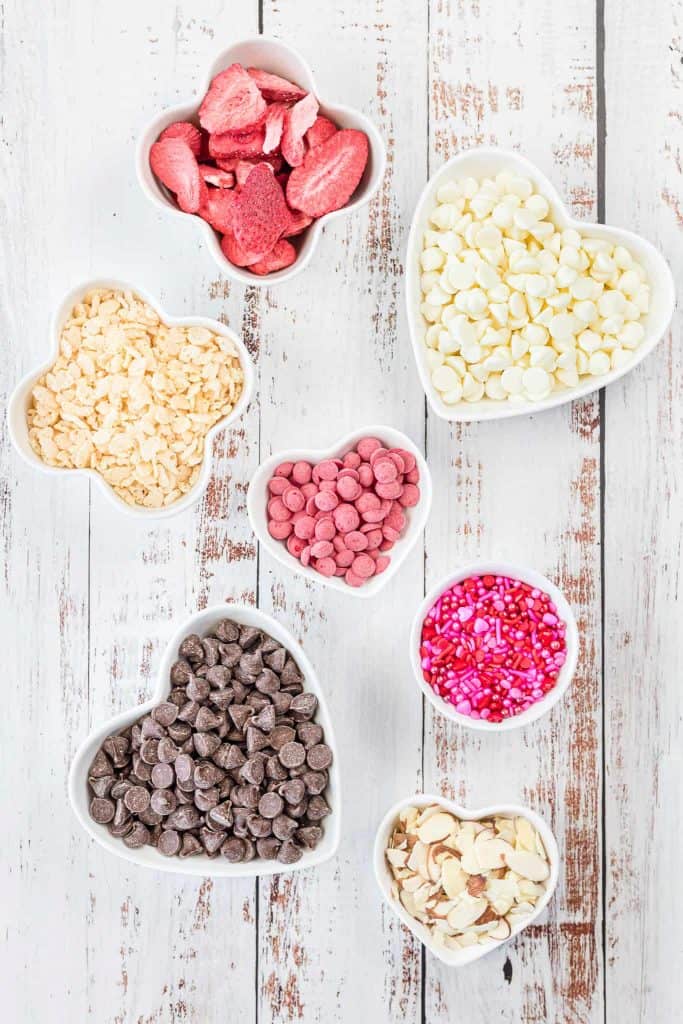 White chocolate chips and milk chocolate chips in white heart bowls. Freeze dried strawberries, pink melting discs, sliced almonds and Valentine's Day sprinkles in white bowls on a white wood background.