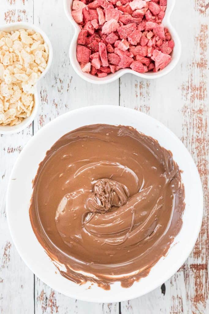Melted milk chocolate in a white bowl with white chocolate chips and freeze dried strawberriesin white bowls above it.