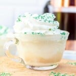 Head on photo of Iced Coffee with whipped cream on top and green sprinkles in a glass mug on a wood background.
