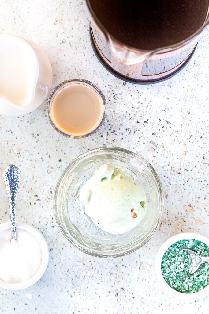 Overhead view of pistachio ice cream in a glass cup with Bailey's liqueur, sugar, cream and green sprinkles in cups around it.
