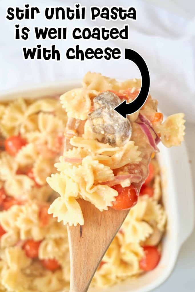 Bow tie pasta coated in cheese on a wooden spoon with baking dish of pasta below it.