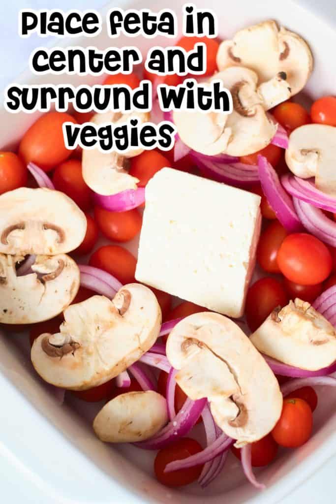 Closeup of feta cheese in a baking dish surrounded by cherry tomatoes, red onions and mushrooms.