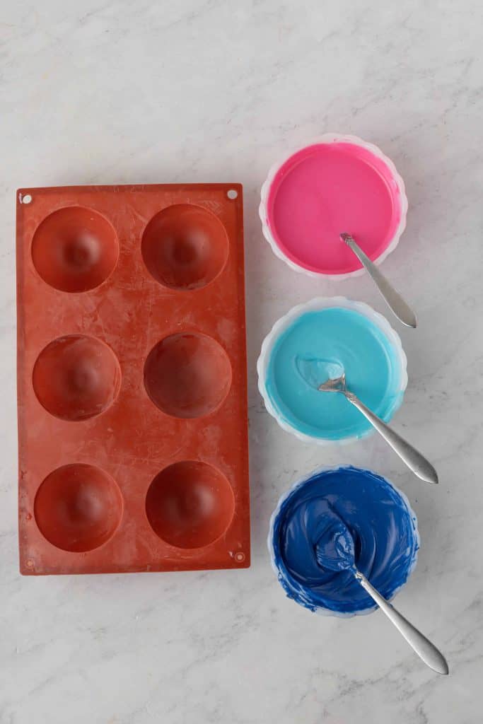 Pink, blue and purple candy melts in white bowls with spoons in them beside a silicone mold.