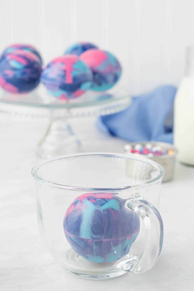 Galaxy hot cocoa bomb in glass cup with 4 bombs in background ion a glass cake stand.