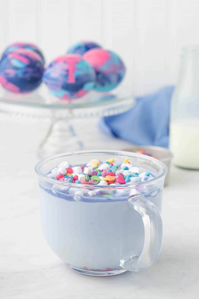 Galaxy Hot Cocoa in a glass cup with hot cocoa bombs on a glass cake stand in the background.