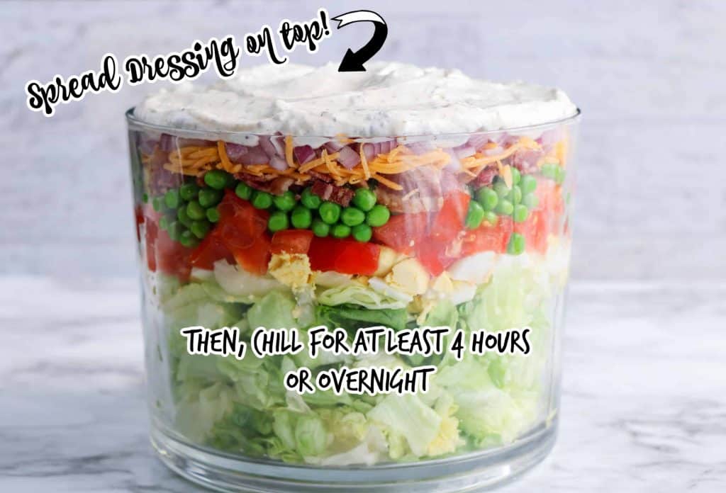 Head on view of layered vegetables in a glass trifle bowl with dressing on top and text overlay of instructions to dress salad and chill!