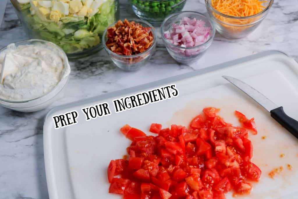 Diced tomatoes on a white cutting board with other ingredients in small bowls behind it on a countertop.