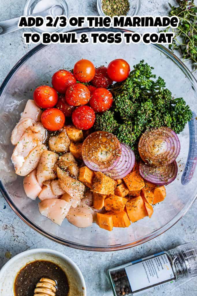Overhead view of uncooked chicken, cherry tomatoes, broccoli, red onion and sweet potatoes with balsamic marinade on top in a glass bowl.
