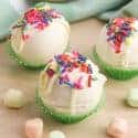Three White Chocolate Hot Chocolate Bombs on a wood counter with rainbow mini marshmallows surrounding them.