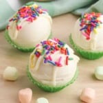 Three White Chocolate Hot Chocolate Bombs on a wood counter with rainbow mini marshmallows surrounding them.