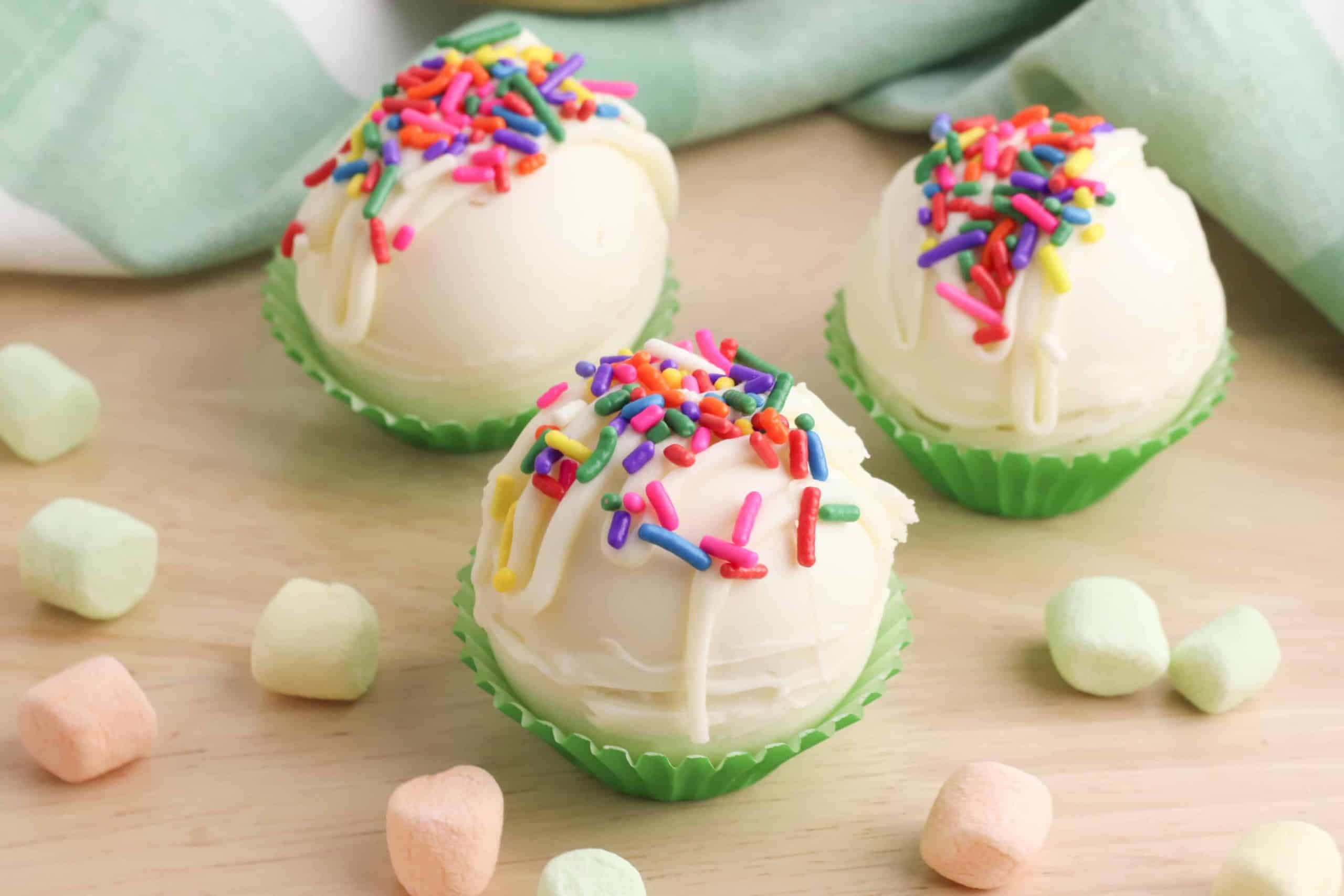 Three White Chocolate Hot Chocolate Bombs on a wood counter with rainbow mini marshmallows surrounding them.