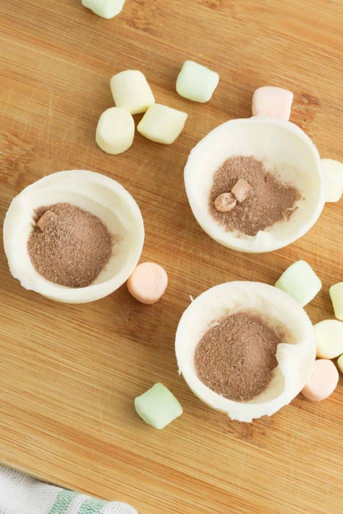 Hot chocolate in half of a white chocolate mold with rainbow marshmallows sprinkled around them on a wood cutting board.