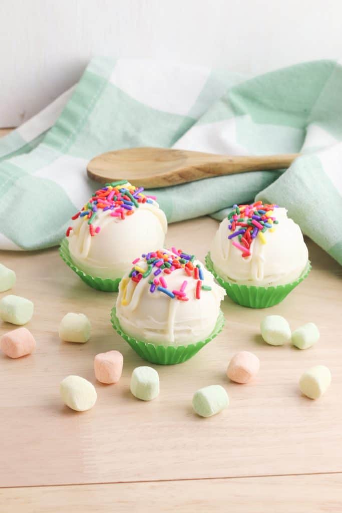 White Chocolate Hot Chocolate Bombs on a wood surface with green and white dish towel with wooden spoon on top in the background.