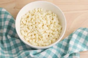 White chocolate chips in a white bowl on a wood background with a light blue and white checked dish towel in the left corner.