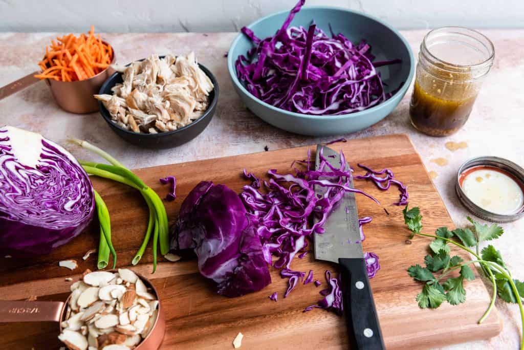 Shredded purple cabbage on a cutting board with green onions, shredded carrots, sliced almonds and cooked chicken on the side.