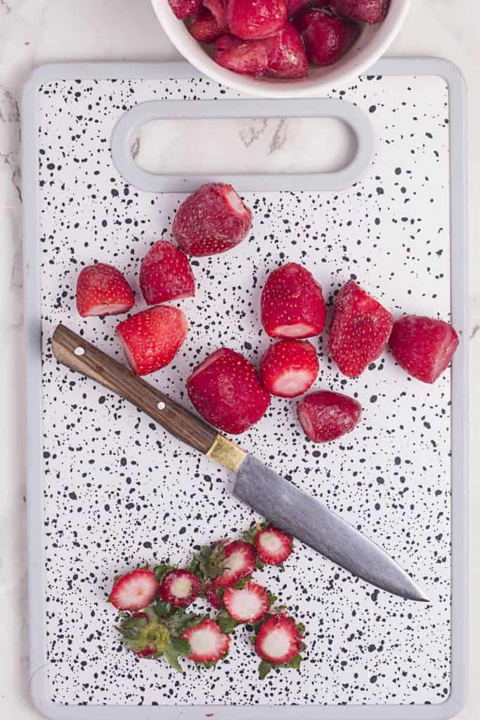 fresh strawberries with tops cut off on a white and black speckled cutting board.
