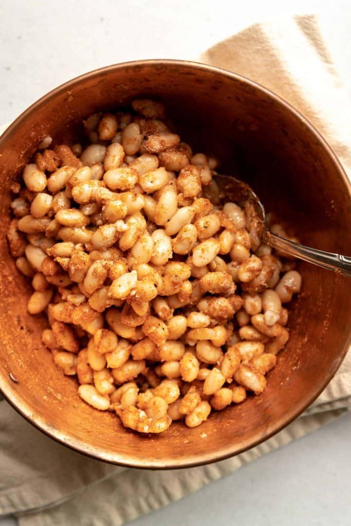 White Beans tossed with spices and olive oil in a brown boil with a spoon in the bowl.