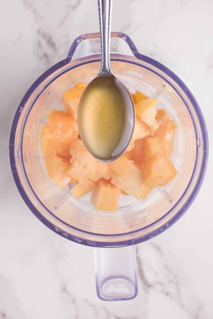 Tablespoon of honey above a blender with frozen mango chunks.