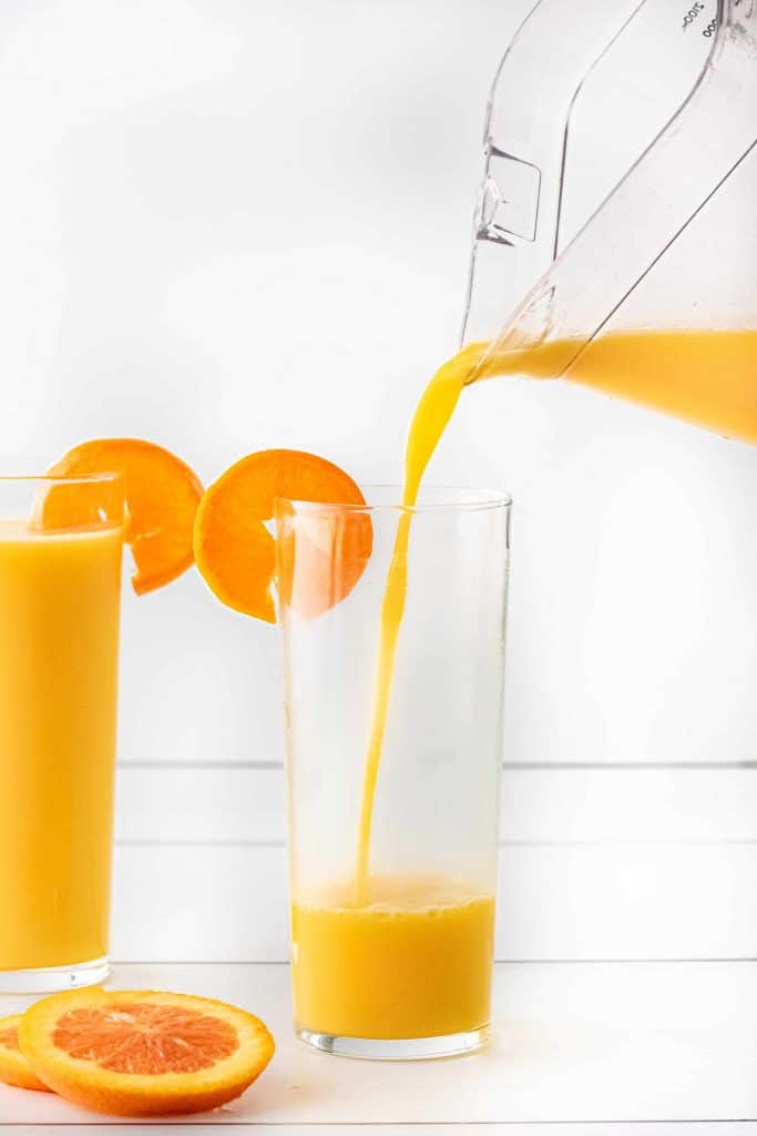 Pouring orange drink into a tall glass with an orange slice garnish on the rim.